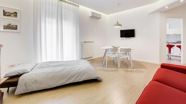 A modern bedroom with a bed, a dining area with a table and chairs, a television mounted on the wall, and a red chair in the foreground. - Lovely 3 Rooms Apartment Close Trastevere Station (Rome)