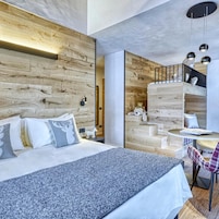 Un dormitorio moderno con cabecera de madera, cama con ropa de cama blanca, una mesita redonda con una silla y una escalera con un osito de peluche.