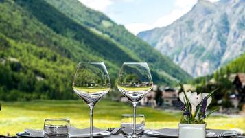 Une table dressée avec deux verres à vin, un verre à eau et un vase rempli de fleurs, le tout mis en valeur par un paysage montagneux en arrière-plan.