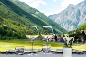 Una mesa servida con dos copas de vino, un vaso de agua y un florero con flores, con montañas al fondo.