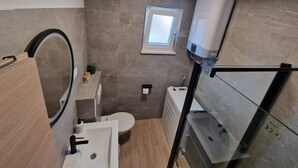 Ein kleines Badezimmer mit Toilette, Waschbecken und Dusche. Ein Fenster sorgt für natürliches Licht, und ein Handtuchhalter ist zu sehen.