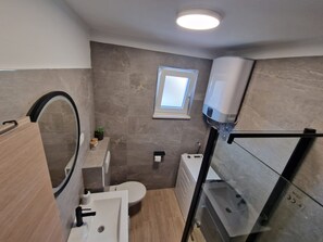 Un bagno compatto con il water, lavandino e doccia. Una finestra permette l'illuminazione naturale, ed è visibile un porta asciugamani.