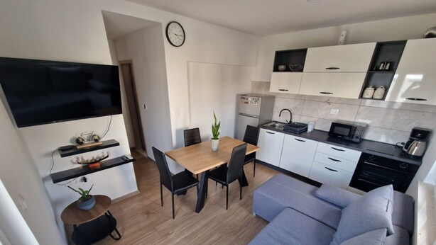 Eine moderne Küche mit weißen Schränken, einer Mikrowelle und einer Kaffeemaschine. Es gibt einen Essbereich mit einem Holztisch und Stühlen, sowie ein graues Sofa im Wohnbereich.