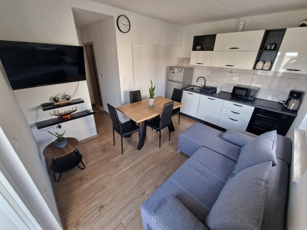 Una cucina moderna con mobili bianchi, un forno a microonde e una macchina da caffè. C'è una zona pranzo con un tavolo e sedie in legno, e un divano grigio nella zona living.