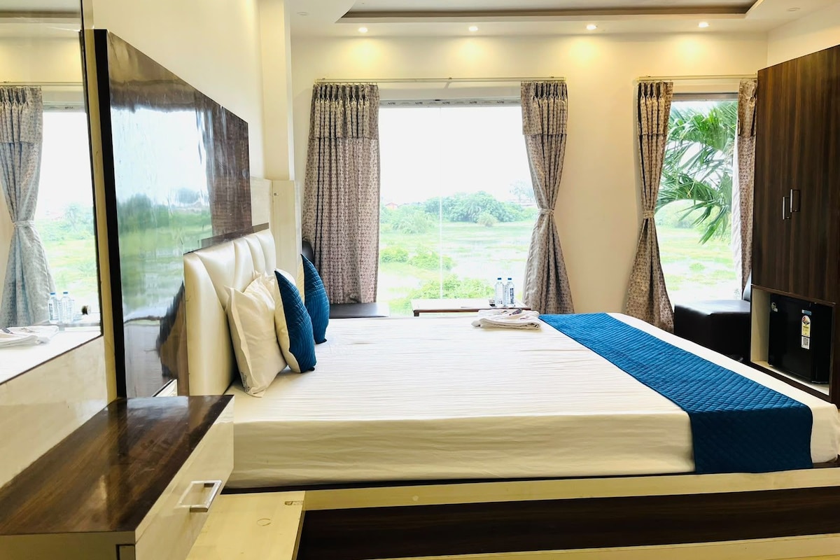 A hotel room with a bed, a flat-screen TV, a window with curtains, and a view of greenery outside.