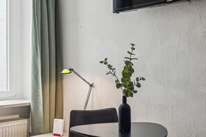 A room with a round table, a black vase with a plant, a black chair, a floor lamp, and a television mounted on the wall. - Centrally Located Apartment in Passau | Washer, Dryer, Wi-Fi, and Pets Welcome (Passau)
