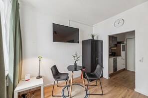 A compact living space with a dining table, two chairs, a floor lamp, a flat-screen TV mounted on the wall, a dark cabinet, and a kitchen area visible through an open door. - Centrally Located Apartment in Passau | Washer, Dryer, Wi-Fi, and Pets Welcome (Passau)