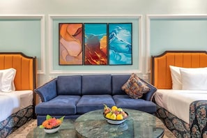 Habitación de hotel con un sofá azul, dos camas con cabeceros naranjas, una mesa de centro redonda con fruta y arte abstracto en la pared.