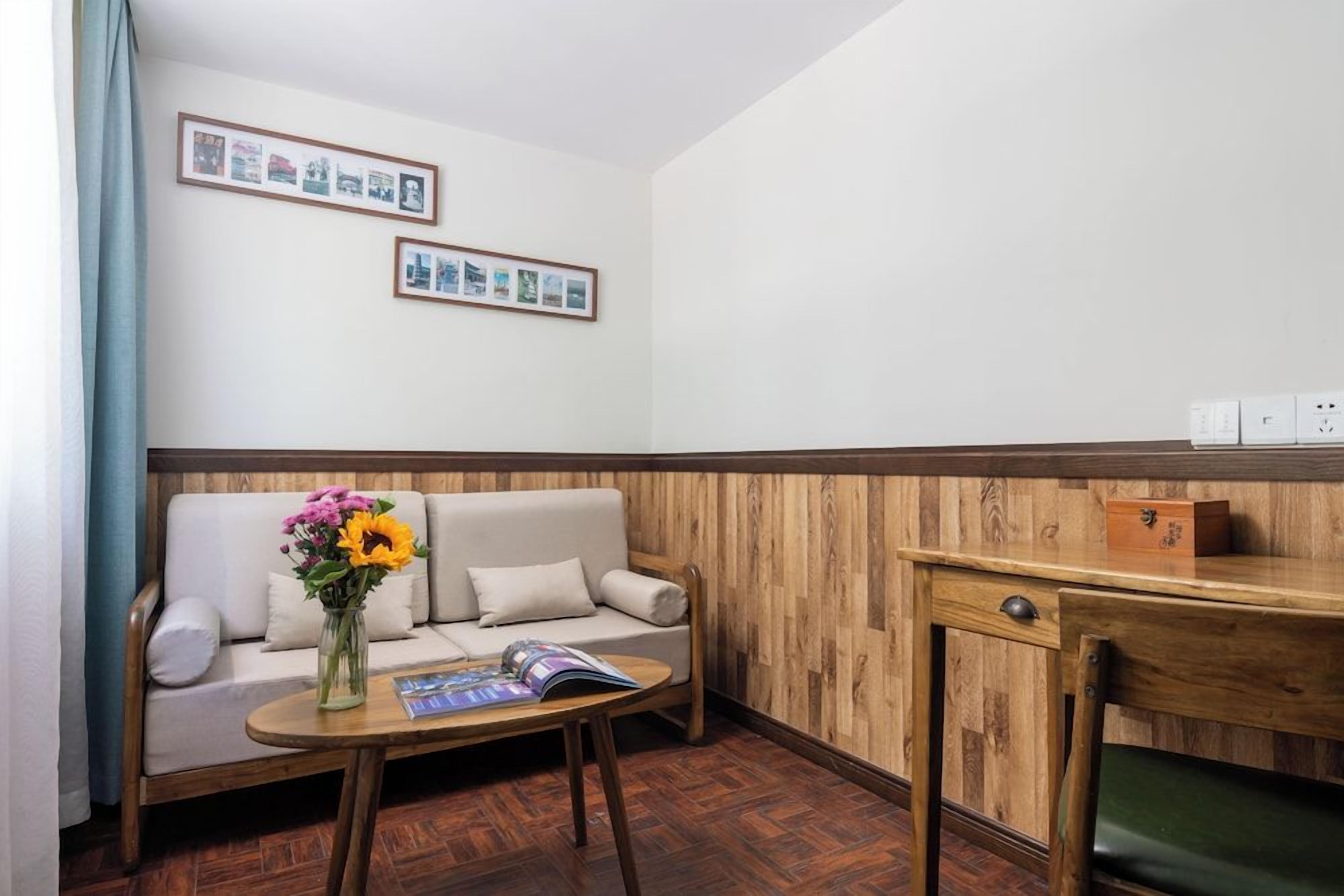 A room with a wooden floor, a sofa with cushions, a small table with a vase of flowers, and a wall with framed pictures.