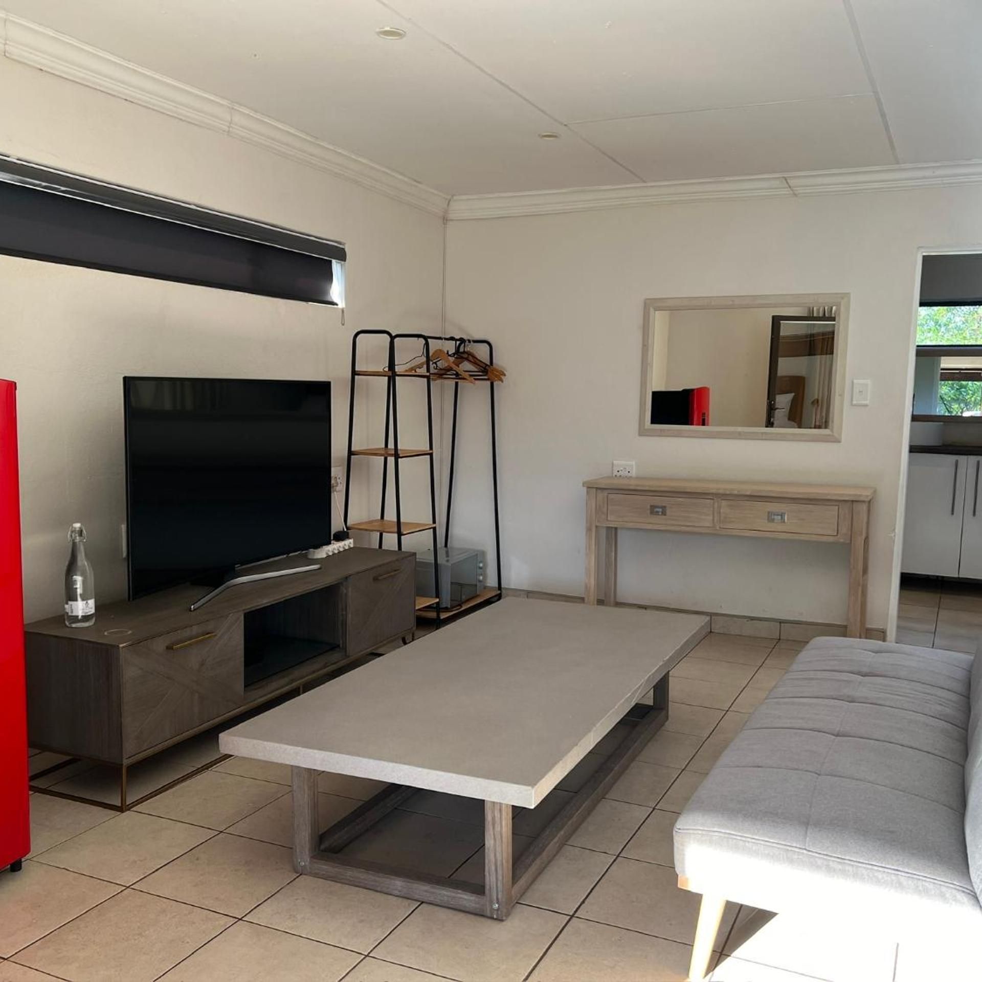 A living room with a flat-screen TV, a coffee table, a sofa, a TV stand, a wooden cabinet, a desk with drawers, a mirror, and a coat rack.