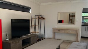 A living room with a flat-screen TV, a coffee table, a sofa, a TV stand, a wooden cabinet, a desk with drawers, a mirror, and a coat rack. - Together Lifestyle Resort (Thulamela)
