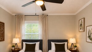 Hotellihuone, jossa on kaksi sänkyä, kattotuuletin, ikkuna verhoilla ja seinään kiinnitetyt lamput.