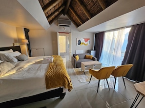 Habitació d'hotel amb llit, escriptori, cadira, taula petita, zona de dutxa i finestra gran amb cortines.