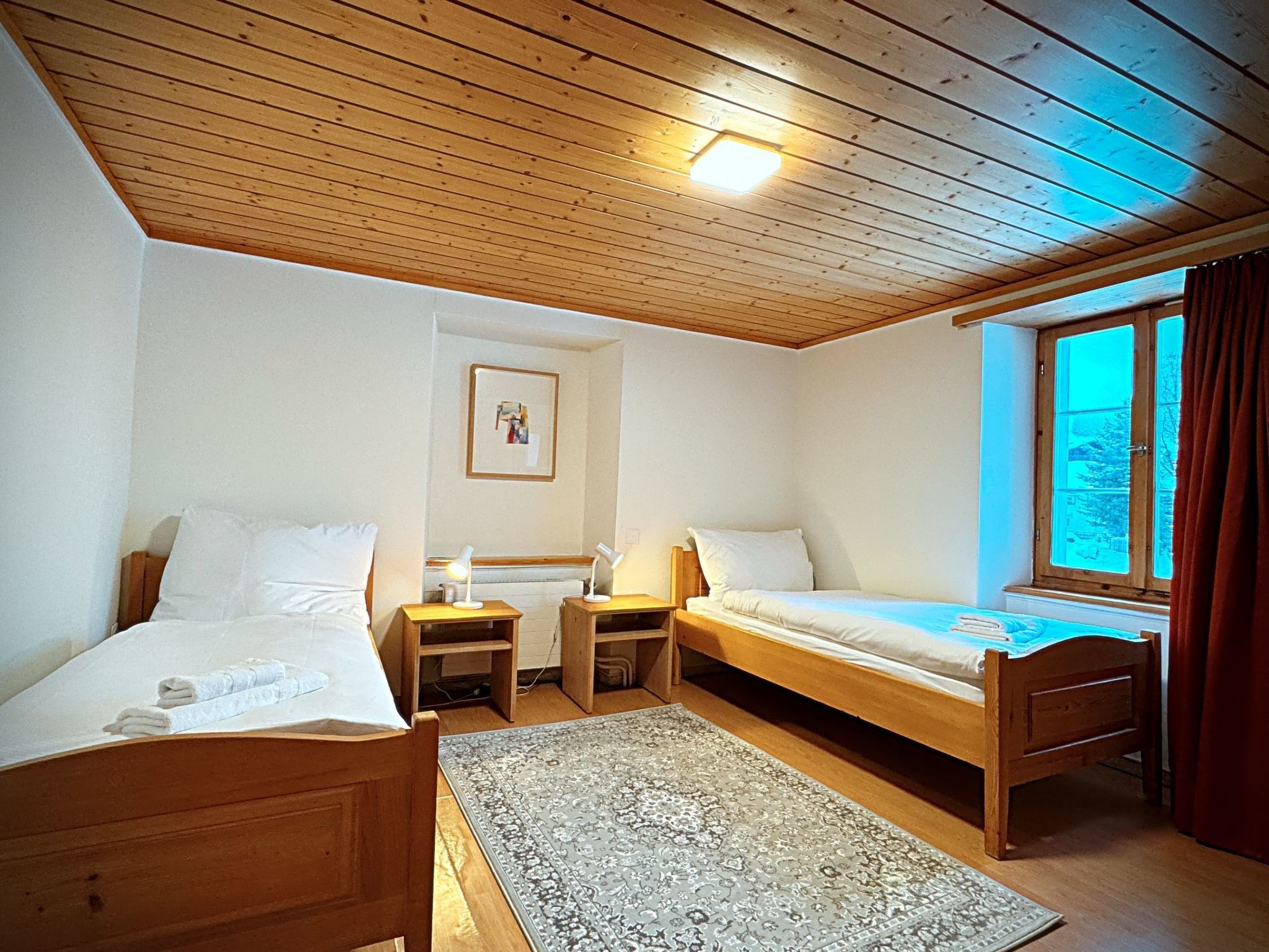 Une chambre avec deux lits en bois, un petit tapis, une table de chevet avec une lampe et une fenêtre avec des rideaux.