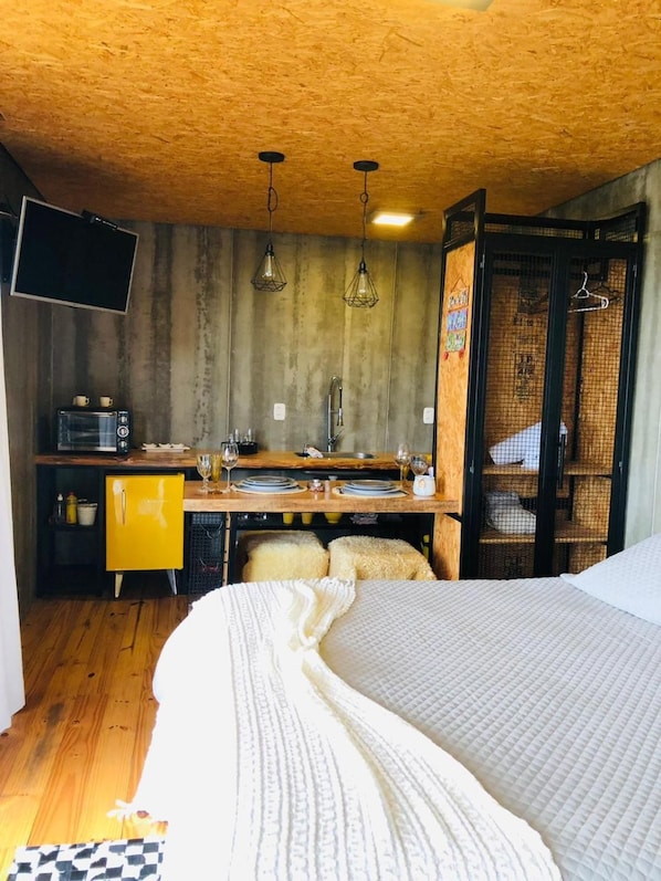 Una habitación con cama, un escritorio de madera con silla, una kitchenette pequeña y un televisor montado en la pared.