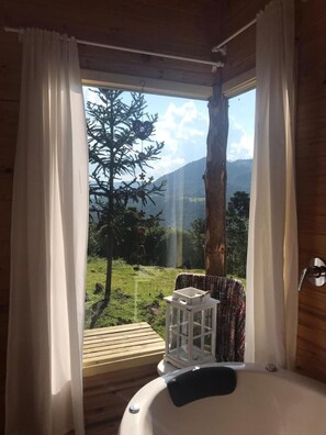 Un baño con bañera, una terraza de madera afuera de la ventana y vista a un árbol y montañas.