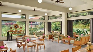 A spacious indoor area with multiple seating arrangements, large windows, ceiling fans, and a view of greenery outside. - Aurika, Coorg - Luxury by Lemon Tree Hotels (Madikeri)