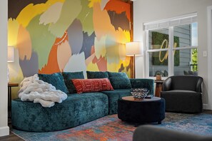 A living room with a teal sofa, a black armchair, a round ottoman, and a colorful abstract painting on the wall. - Walk-In 3BR | Fun Bunk Room + Arcade | Sleeps 9 (Branson)