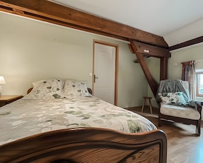 Ein Schlafzimmer mit einem Bett, einem Sessel, einer Holzbalkendecke, einer Tür und einem Fenster mit Vorhängen.