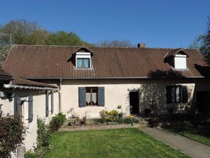 Ein zweistöckiges Haus mit einem Ziegeldach, einer kleinen Veranda mit Stühlen und einem Garten mit verschiedenen Pflanzen.
