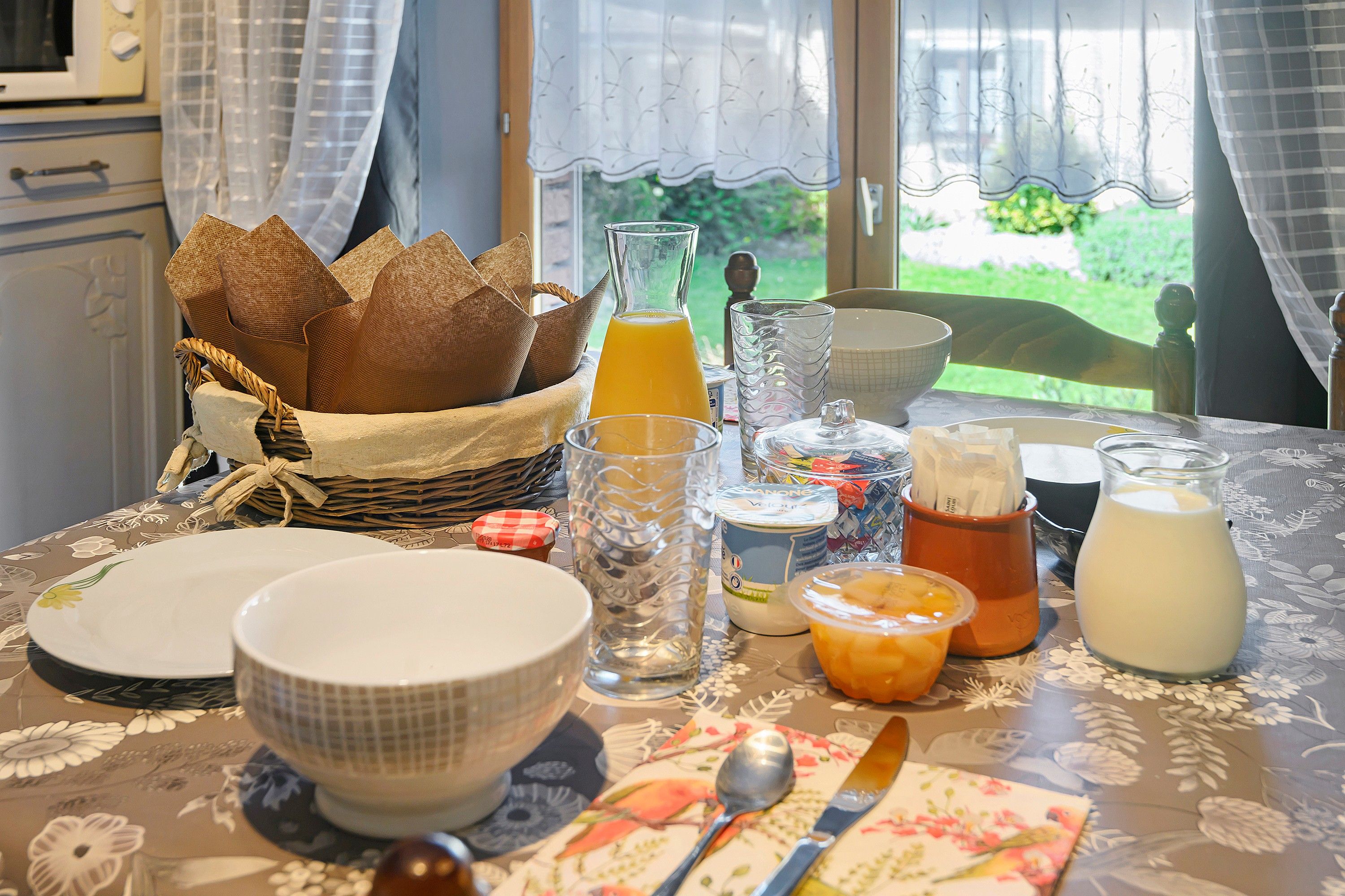 Une table de petit-déjeuner dressée avec différents plats, notamment du pain, un verre de jus d’orange, un bol de céréales et une carafe de lait.