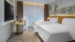 Ein Hotelzimmer mit zwei Betten, einem Schreibtisch mit Laptop, einem Fernseher und einem Landschaftsbild.