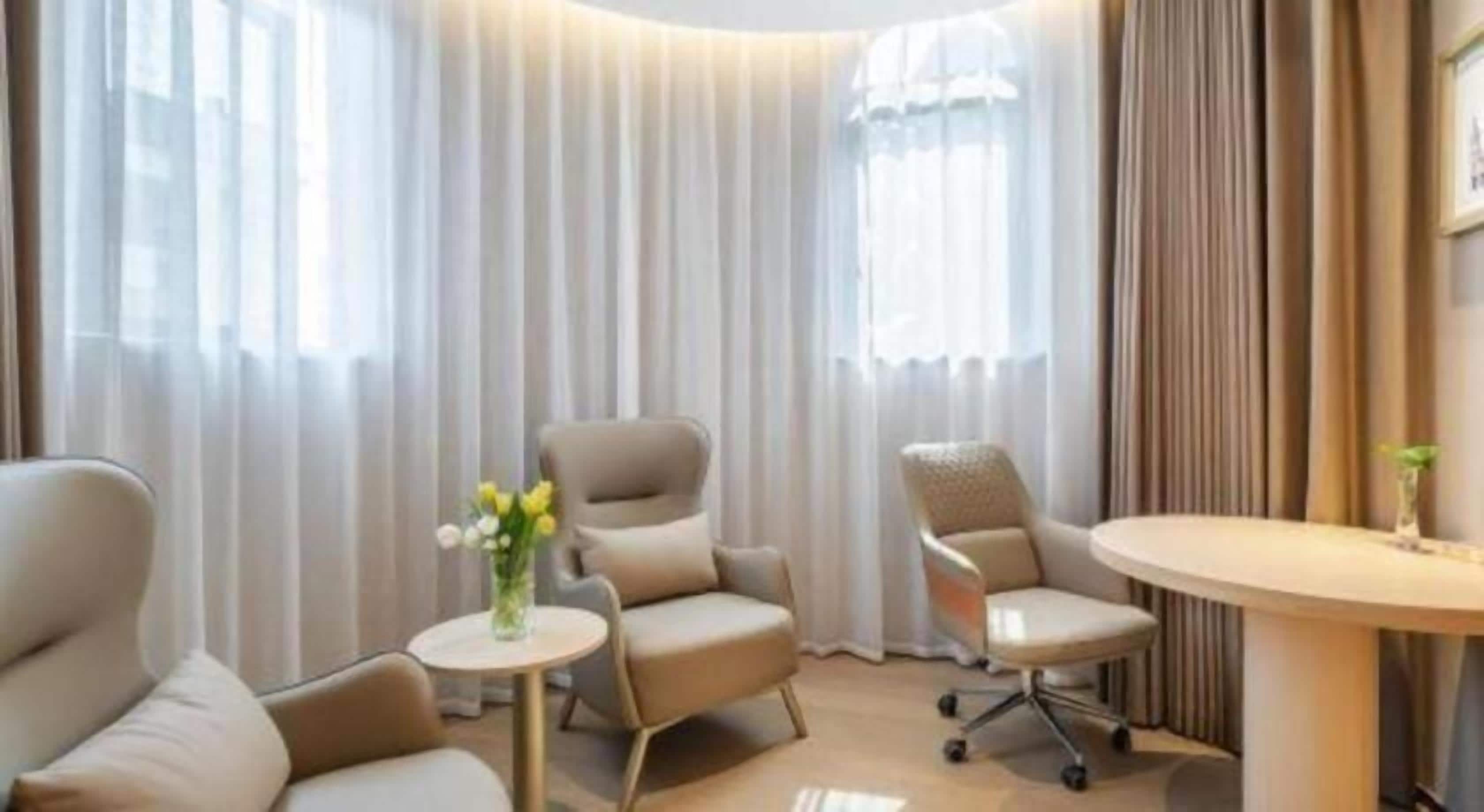 Quarto de hotel com área de estar, incluindo duas poltronas e uma pequena mesa redonda, um vaso com flores e uma janela grande com cortinas.