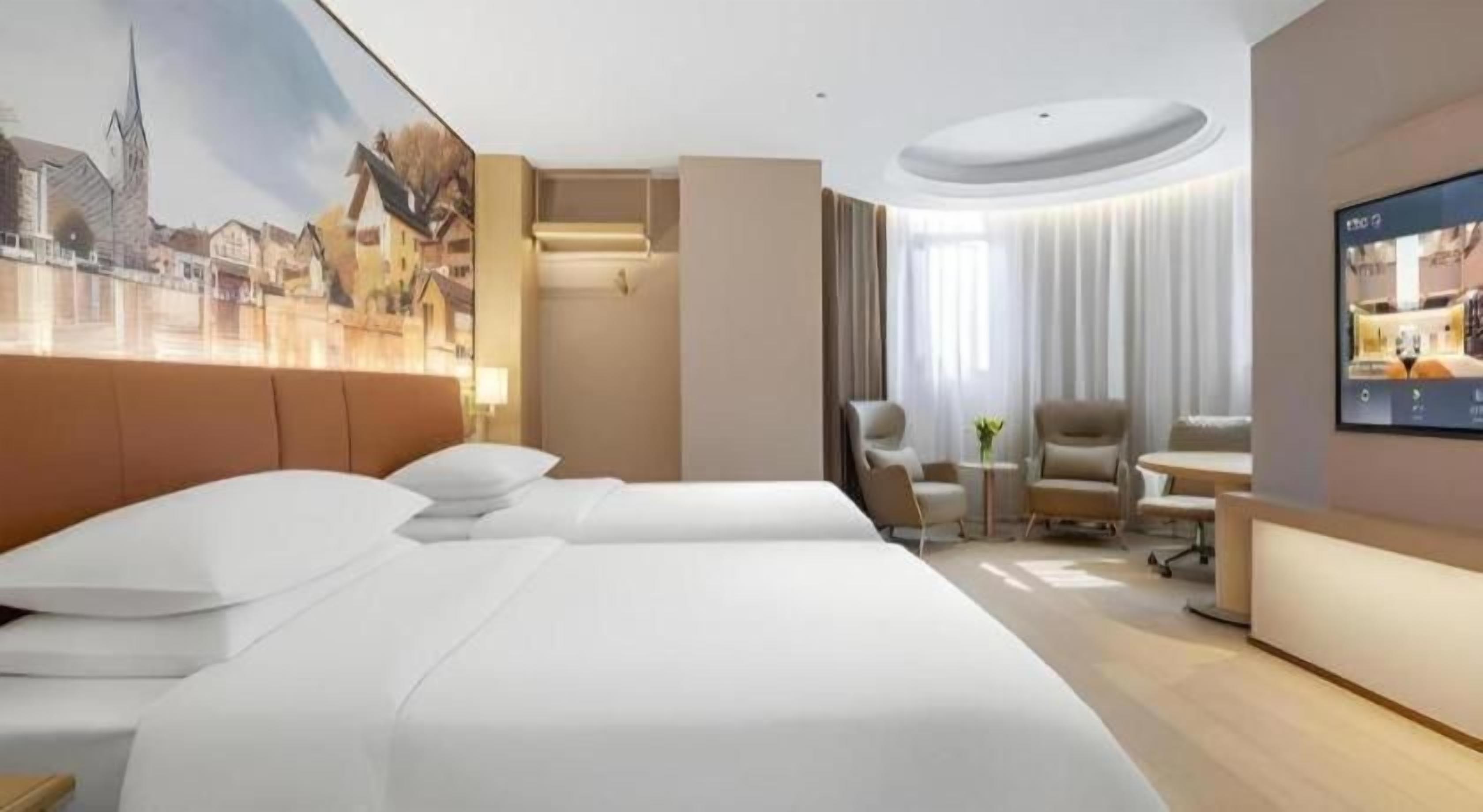 Quarto de hotel com uma cama grande, escrivaninha, duas cadeiras, uma mesa pequena e uma televisão.