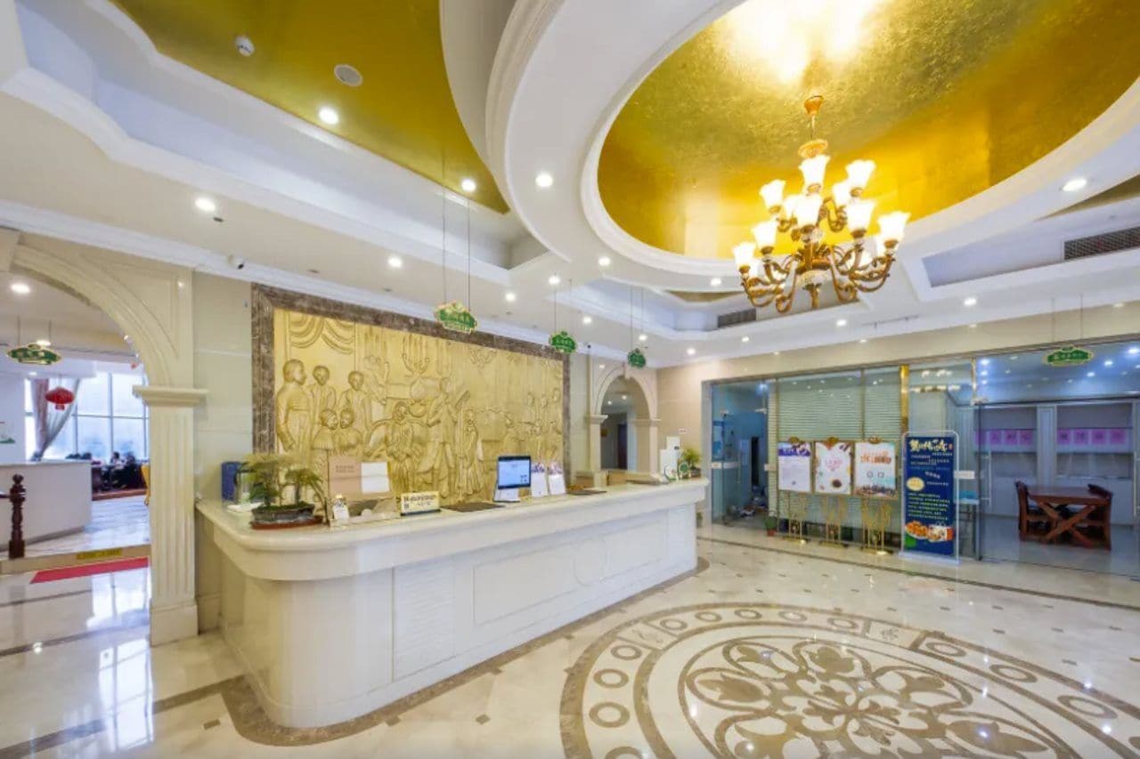 Uma área de recepção de hotel com piso de mármore, balcão de atendimento, lustre e um painel de relevo na parede.