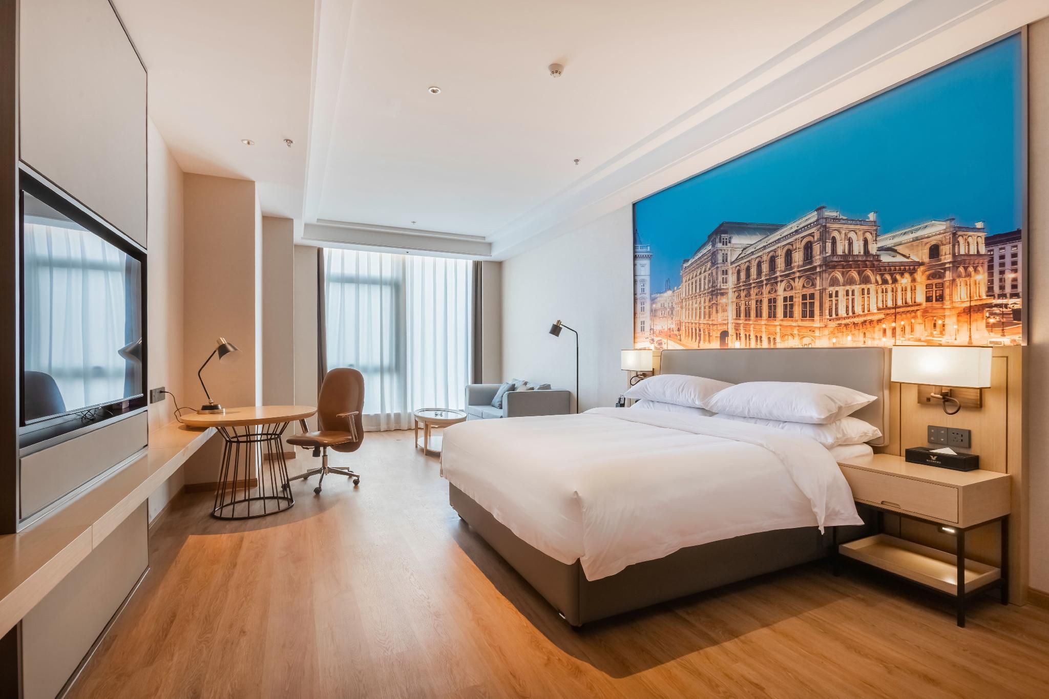 A modern hotel room with a large bed, a desk, a chair, a TV, and a cityscape mural on the wall.