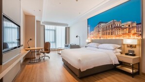 Ett modernt hotellrum med en stor säng, ett skrivbord, en stol, en TV och en väggmålning som föreställer en stadsbild.