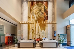 Zona de recepción de un hotel con paredes de mármol, un mural grande, dos mostradores de recepción y una pantalla digital.