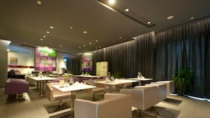 En moderne hotelrestaurant med flere borde og stole, et centralt barområde, og et stort vindue med gardiner.