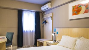 Une chambre d’hôtel comprenant un lit, un bureau, une chaise, un appareil de climatisation, un tableau et une fenêtre avec des rideaux.