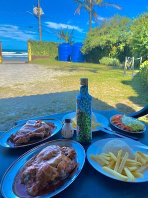 Un repas servi sur une table à l’extérieur, avec vue sur l’océan et la verdure.