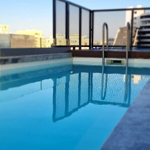 A rooftop swimming pool with clear blue water, metal railings, and a cityscape in the background. - Eleven Apartments (Sliema)