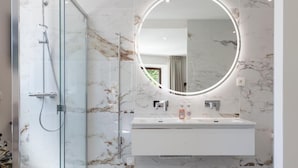 A modern bathroom with a glass shower enclosure, a marble-tiled wall, a round mirror with a light, and a white sink with two faucets. - La Bastide Bourrelly - Mathias Dandine (Cabries)