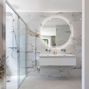 A modern bathroom with a glass shower enclosure, a marble-tiled wall, a round mirror with a light, and a white sink with two faucets. - La Bastide Bourrelly - Mathias Dandine (Cabries)