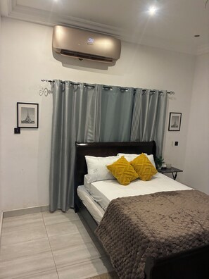 A bedroom with a bed, a nightstand, a wall-mounted air conditioner, and two framed pictures. -  Heritage Comfort & Peace, Your Perfect Getaway. Self compound, gated house (Accra)