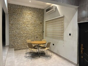A room with a stone wall, a round table with chairs, a window with blinds, and a door with a handle. -  Heritage Comfort & Peace, Your Perfect Getaway. Self compound, gated house (Accra)