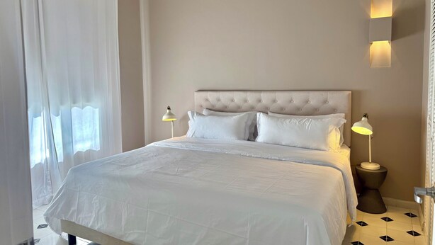 Habitación de hotel con una cama grande, dos lámparas de noche, una luz en la pared y un ventanal con cortinas.