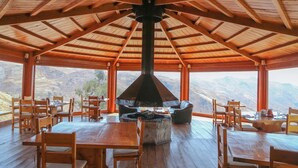 Un'area pranzo coperta con tavoli e sedie in legno, un focolare centrale e vista sulle montagne.