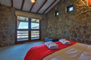 A room with stone walls, a bed with red and white linens, a window with a view, and a small window on the wall.