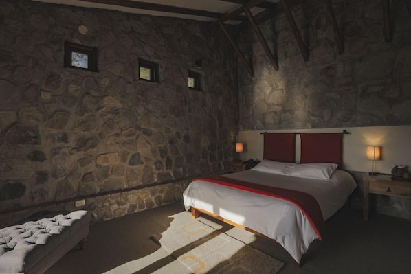 A bedroom with a stone wall, a bed with white bedding and red headboard, a nightstand with a lamp, and a small window.
