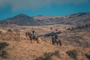 Two individuals on horseback in a dry, hilly landscape with mountains in the background.