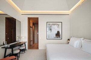 A modern hotel room with a bed, a desk with two chairs, a large abstract painting, and a wooden wardrobe.