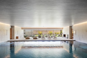 A modern indoor swimming pool with a clear view of the outdoors, surrounded by lounge chairs and a brick wall.