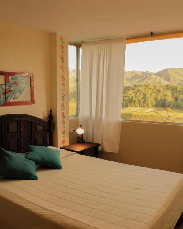 A bedroom with a bed, a nightstand, a lamp, a window with curtains, and a painting on the wall. - Casa Encuentro Ecolodge (Guatapé)