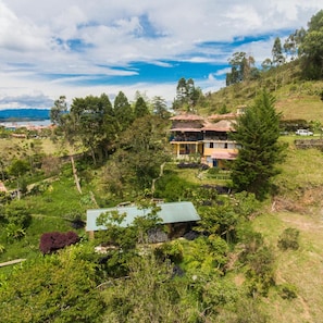 Uma propriedade numa encosta com uma casa de dois andares, com varanda, rodeada por vegetação exuberante e vista ampla da paisagem.