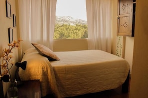 Quarto com cama, janela com cortinas e vista para árvores e montanhas.
